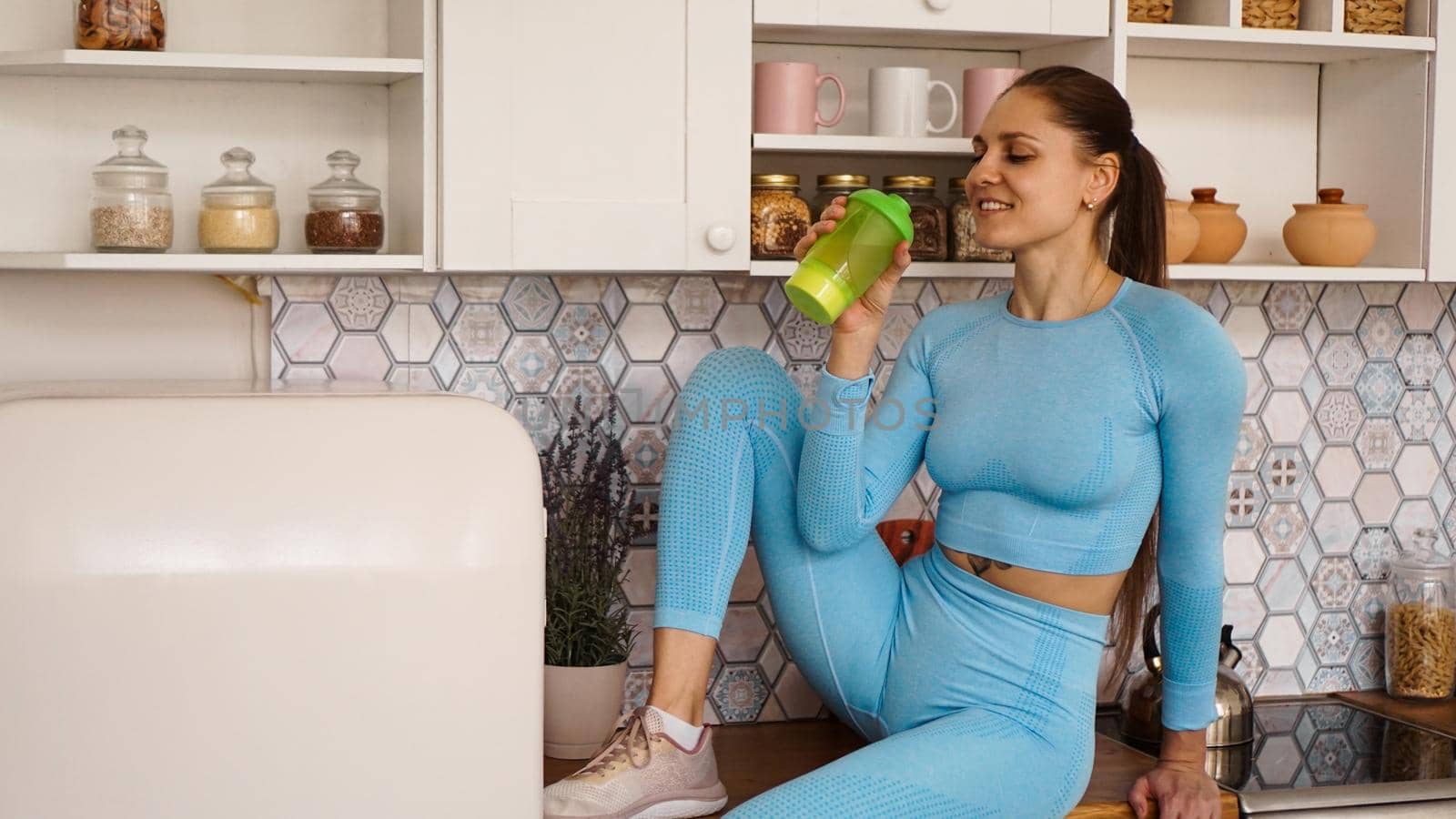 A woman sits on the countertop in the kitchen with a green bottle for sports nutrition or water. Healthy lifestyle concept.