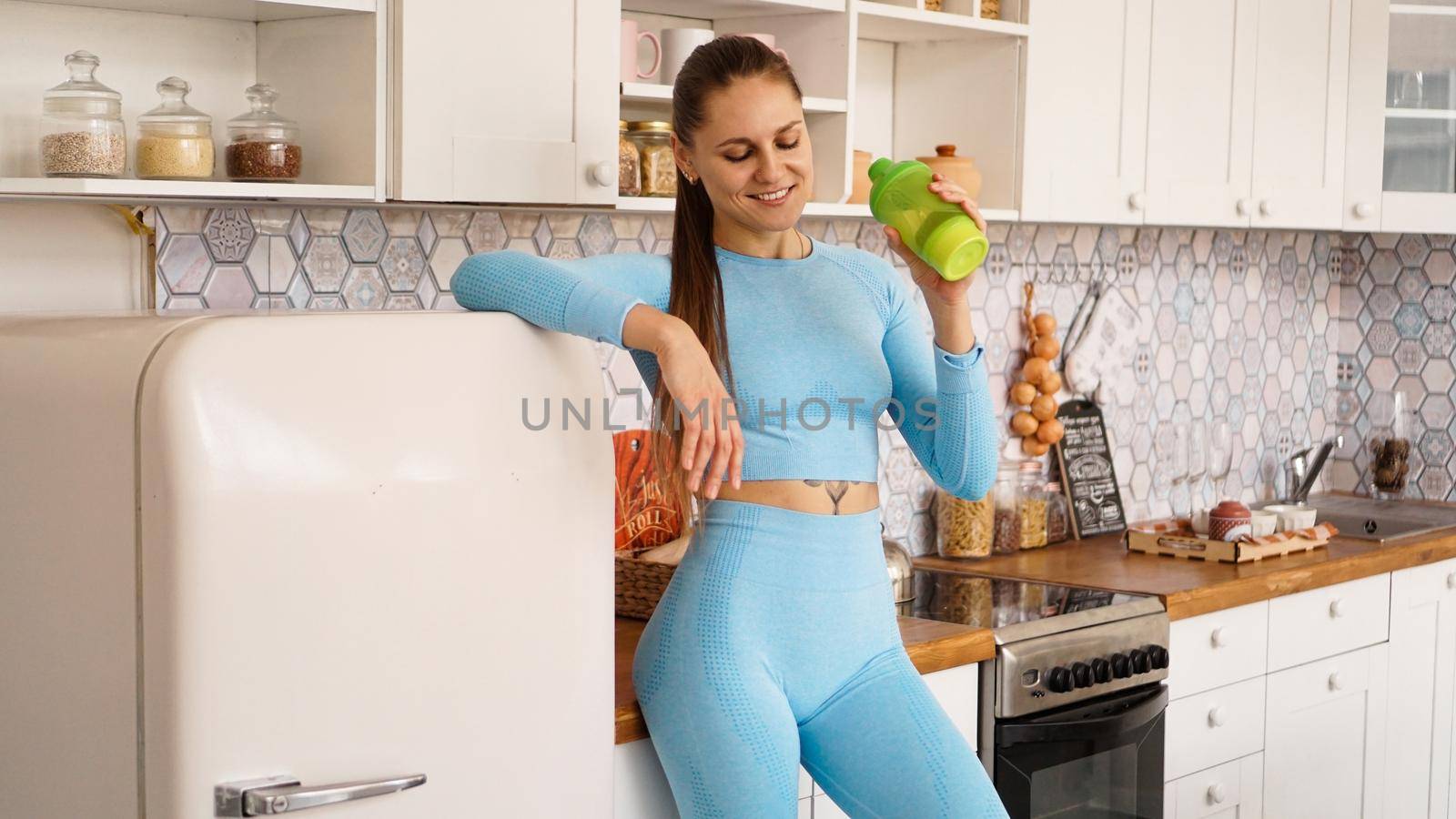 Healthcare and diet concept. Beautiful woman drinks water from a sports bottle after a workout at home. She is in the kitchen near the refrigerator.