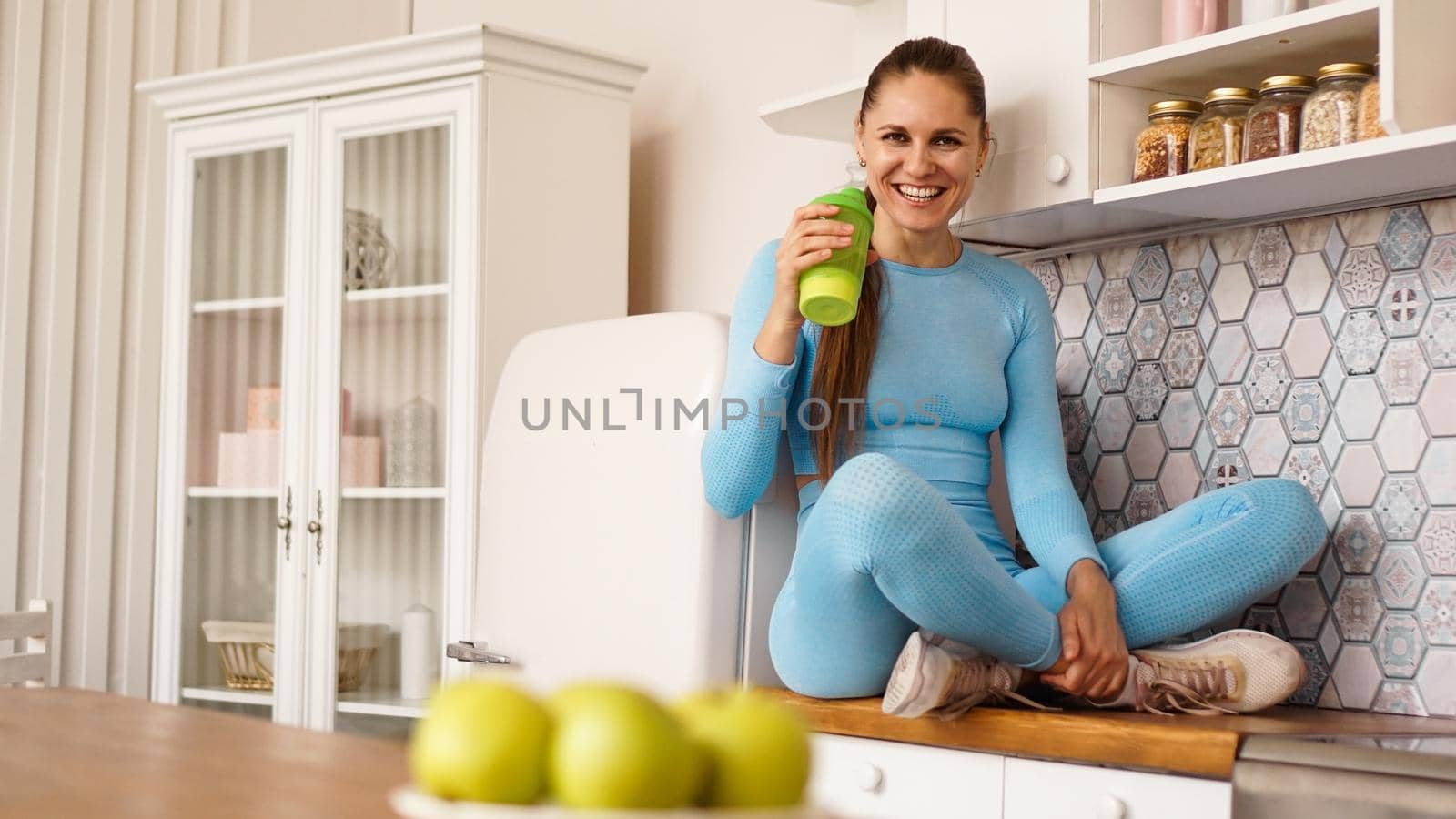 A woman sits on the countertop in the kitchen with a green bottle for sports nutrition or water. Healthy lifestyle concept.