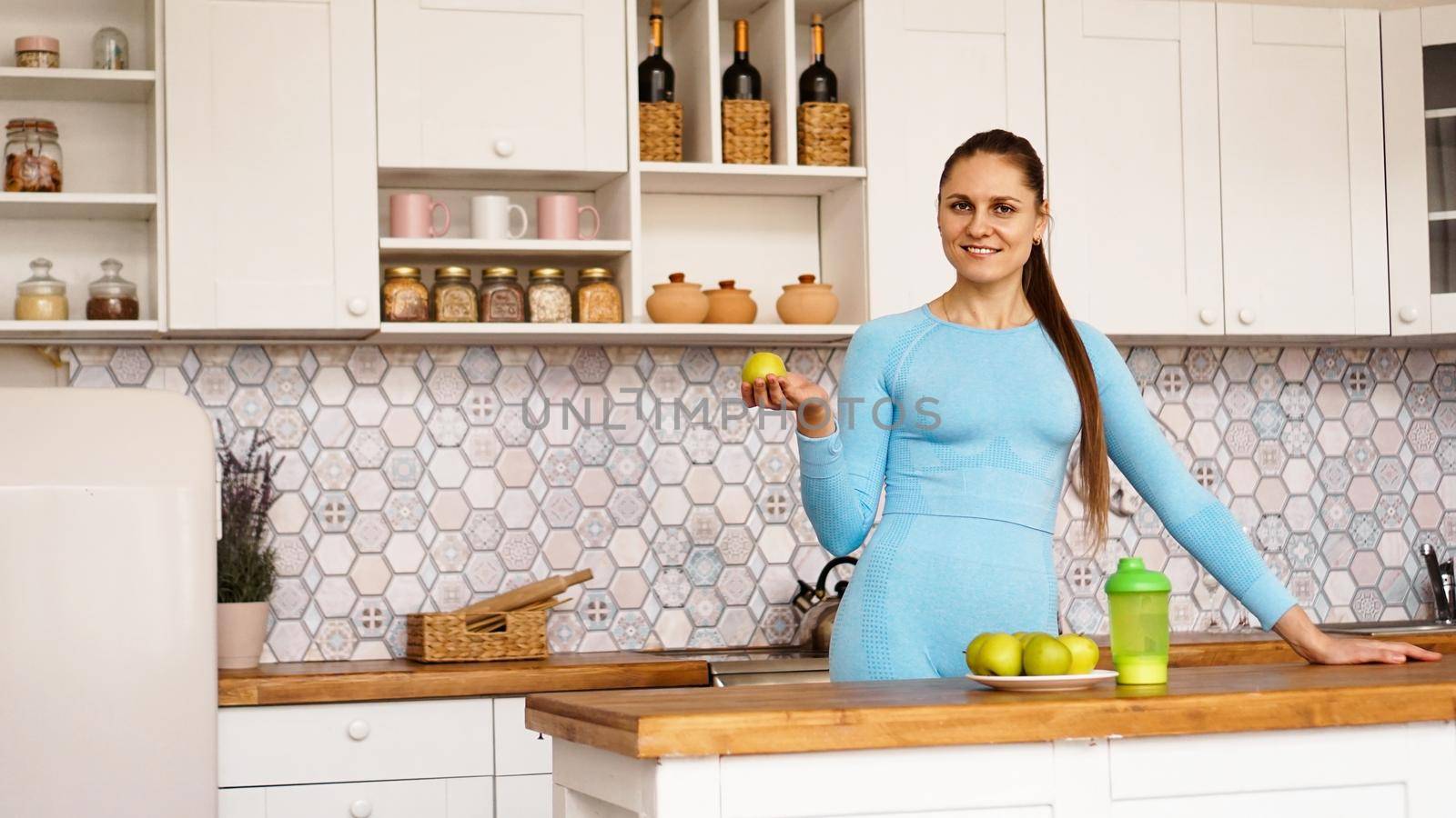 A beautiful woman in a blue suit is standing in the kitchen, smiling and holding a green apple. The concept of sports and weight loss at home.