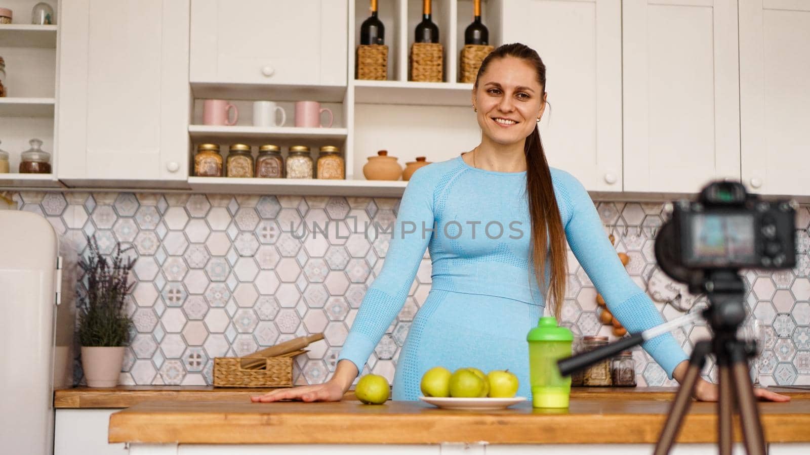 Satisfied healthy young girl recording her video blog episode about healthy food while standing at the kitchen at home. Woman is friendly and smiling