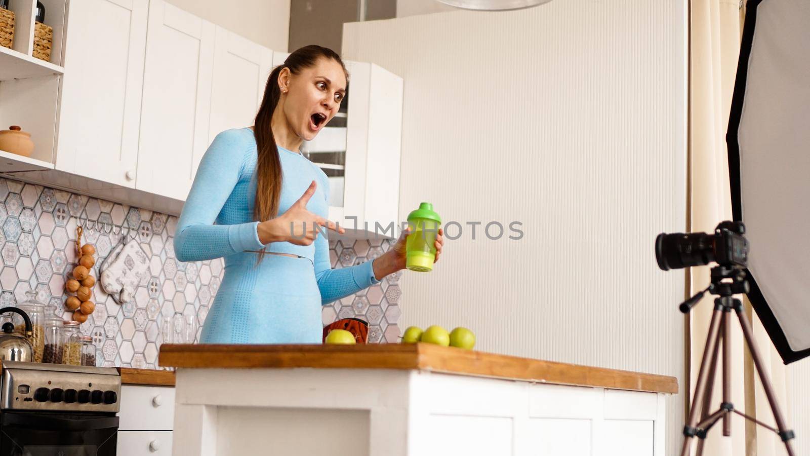 Healthy woman recording her video blog about healthy food additives while standing at kitchen. She is holding a bottle of sports nutrition and smiling