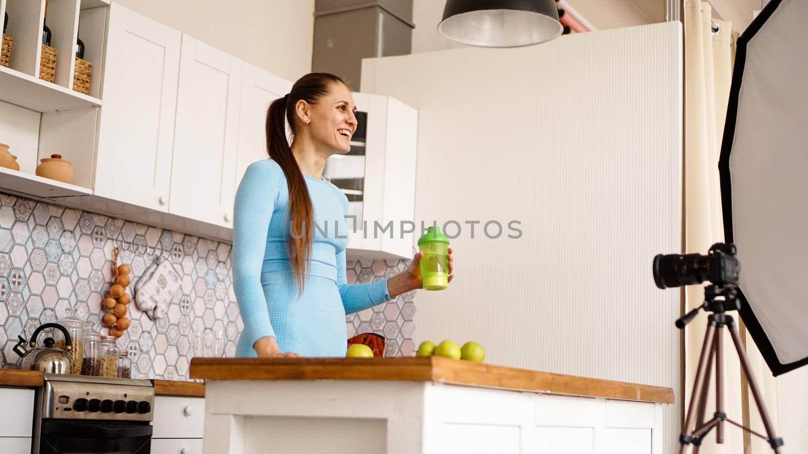 Healthy woman recording her video blog about healthy food additives while standing at kitchen. She is holding a bottle of sports nutrition and smiling