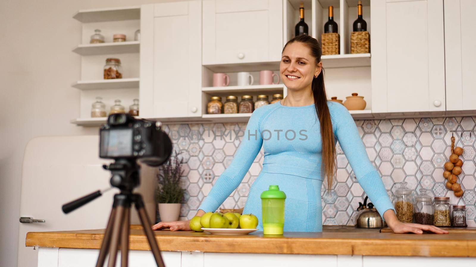 Satisfied healthy young girl recording her video blog episode about healthy food while standing at the kitchen at home. Woman is friendly and smiling