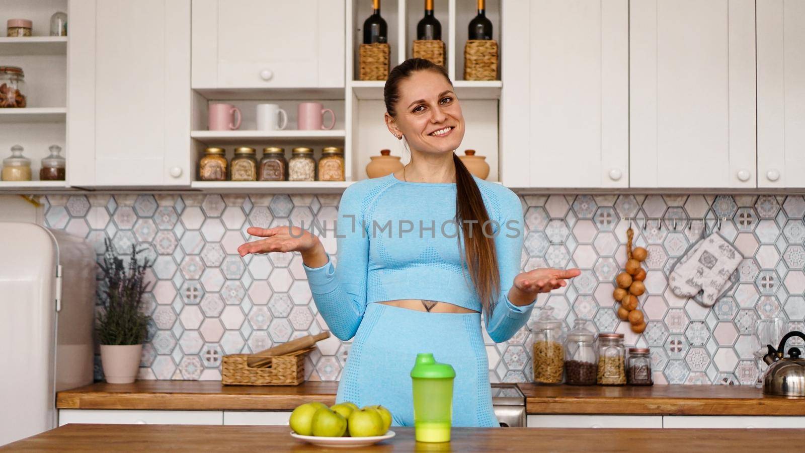 Athletic woman talks about healthy eating in the kitchen and laughs. On the table are green apples and a green bottle for a sports drink and water