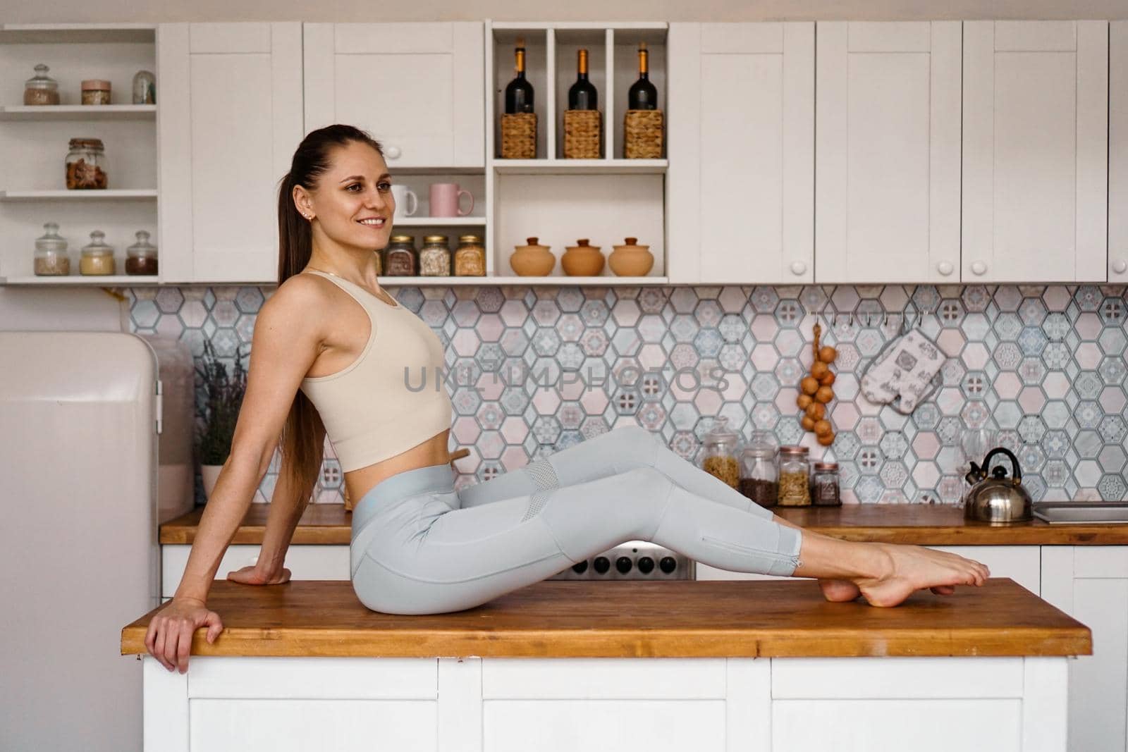 Athletic woman in sportswear posing on the table top of a light modern kitchen. The concept of beauty, health, proper nutrition.