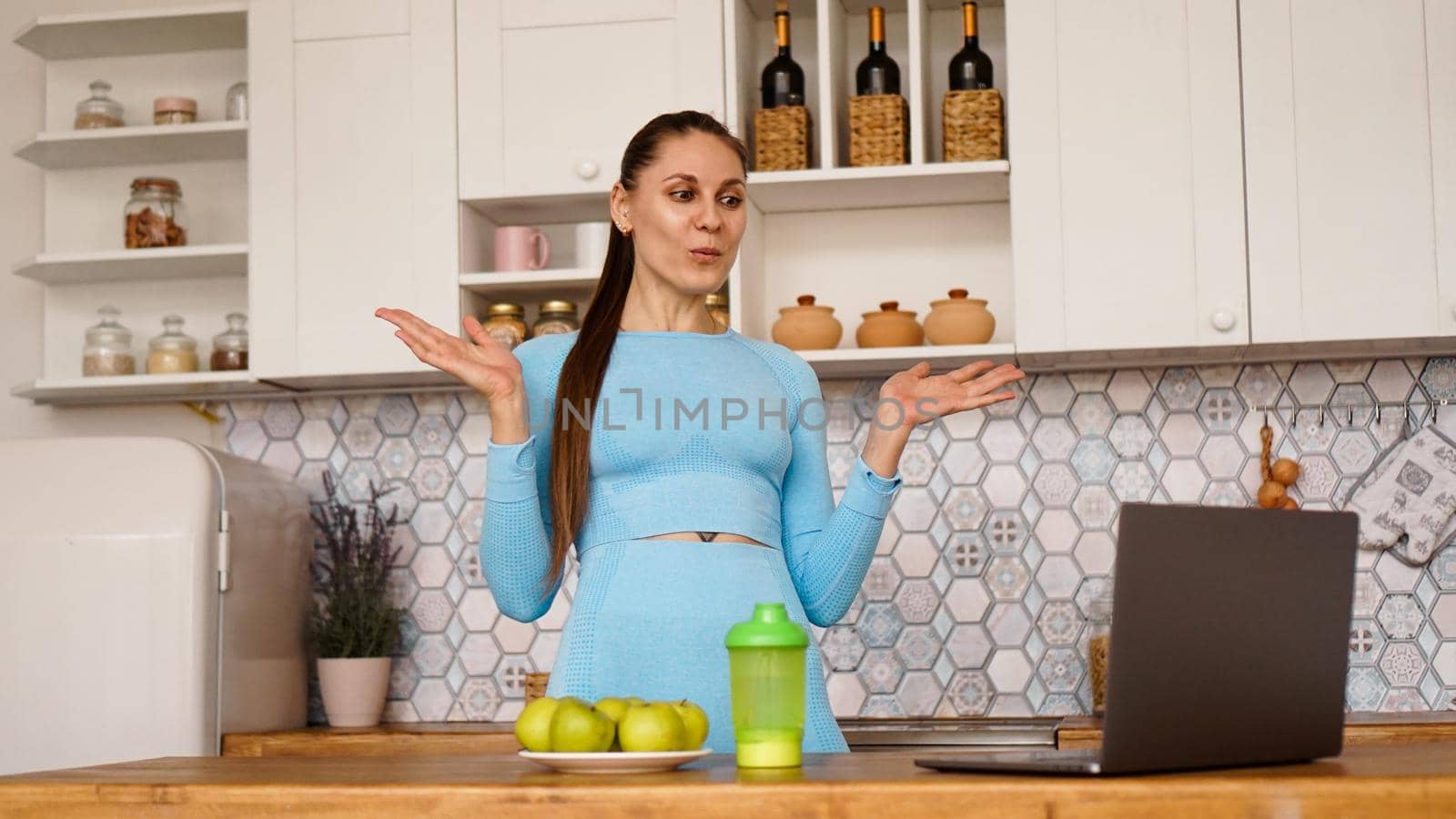 Smiling woman using computer in modern kitchen interior. Cooking and healthy lifestyle concept. Woman broadcasts online and gestures emotionally