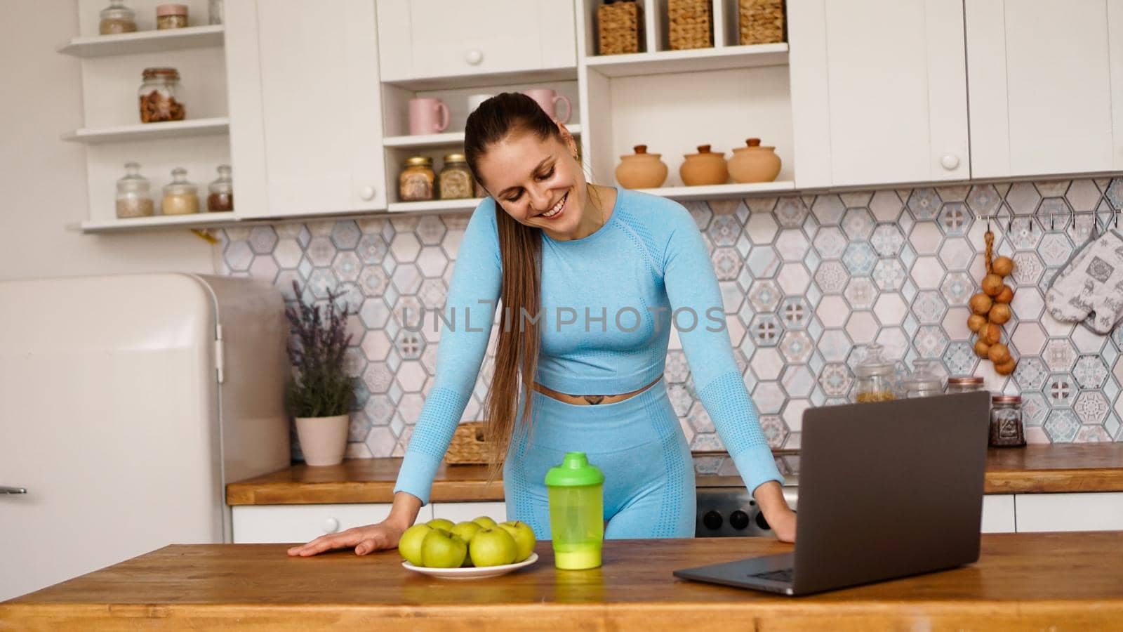 Smiling woman using computer in modern kitchen interior. Cooking and healthy lifestyle concept. Woman broadcasts online and gestures emotionally