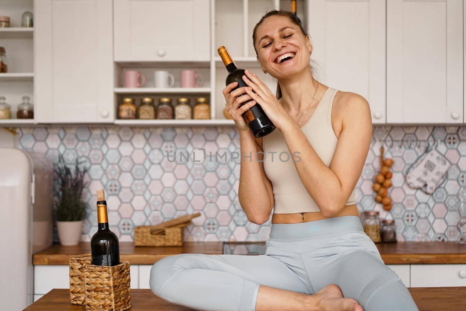 Athletic woman in a tracksuit in a light kitchen drinks red wine from a bottle after doing sports. Woman laughs