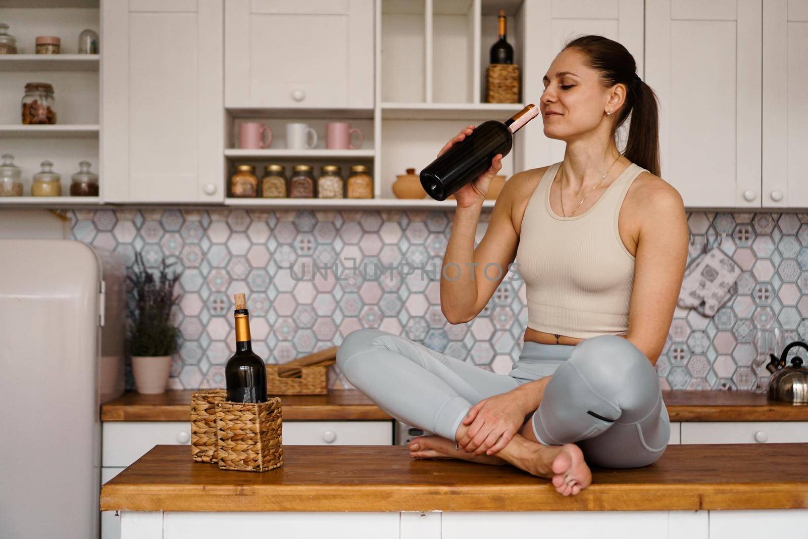 Athletic woman in a tracksuit in a light kitchen drinks red wine from a bottle by natali_brill