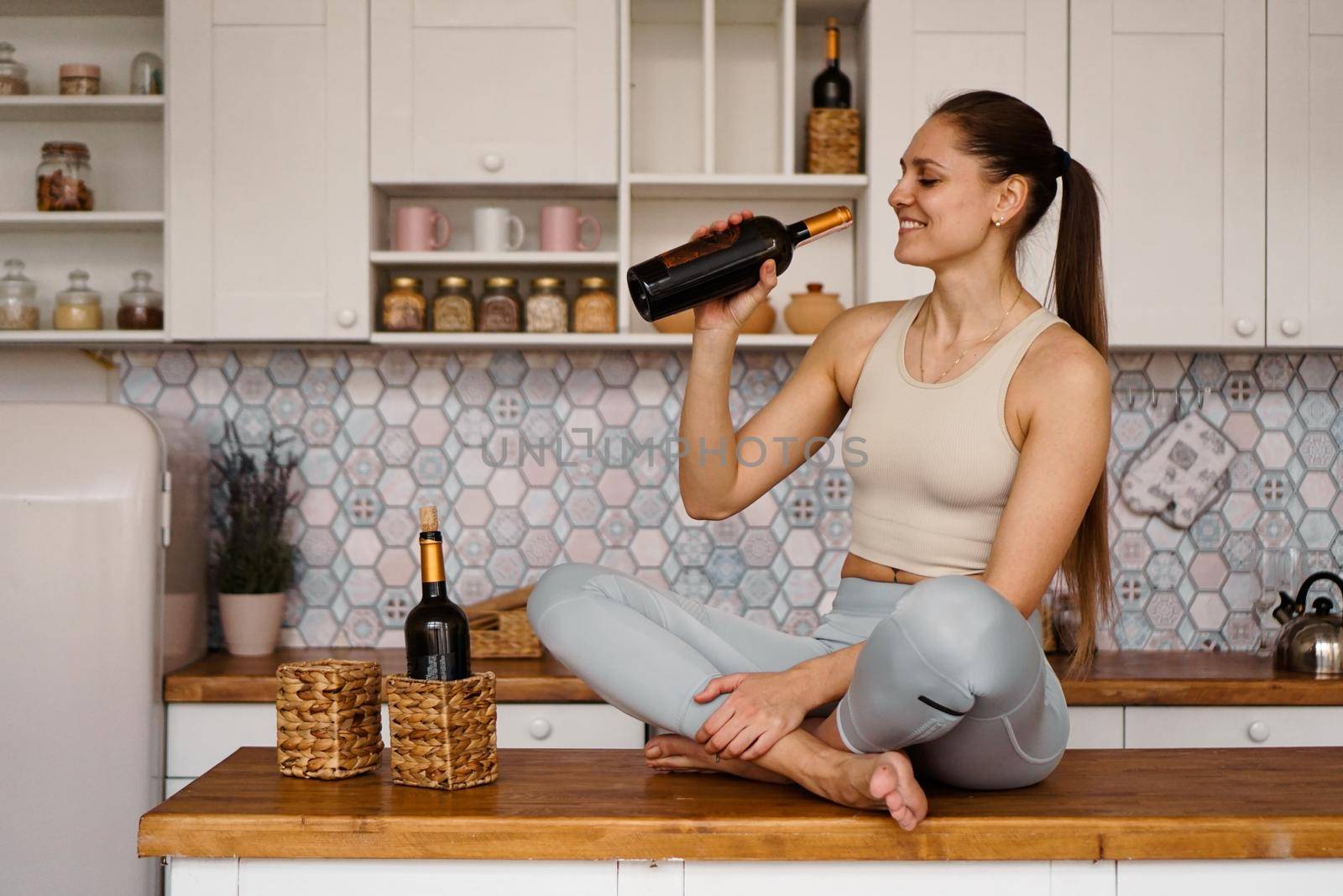 Athletic woman in a tracksuit in a light kitchen drinks red wine from a bottle after doing sports.