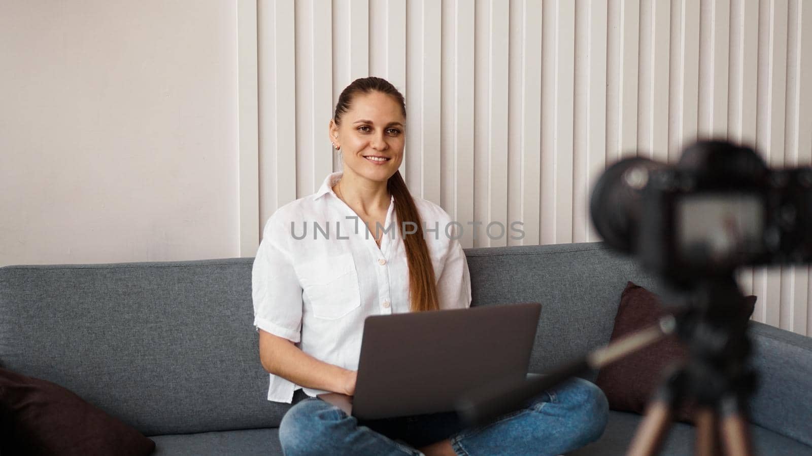 Smiling female blogger records a new video. She sits on the couch at home and holds a laptop. Positive business blog concept.