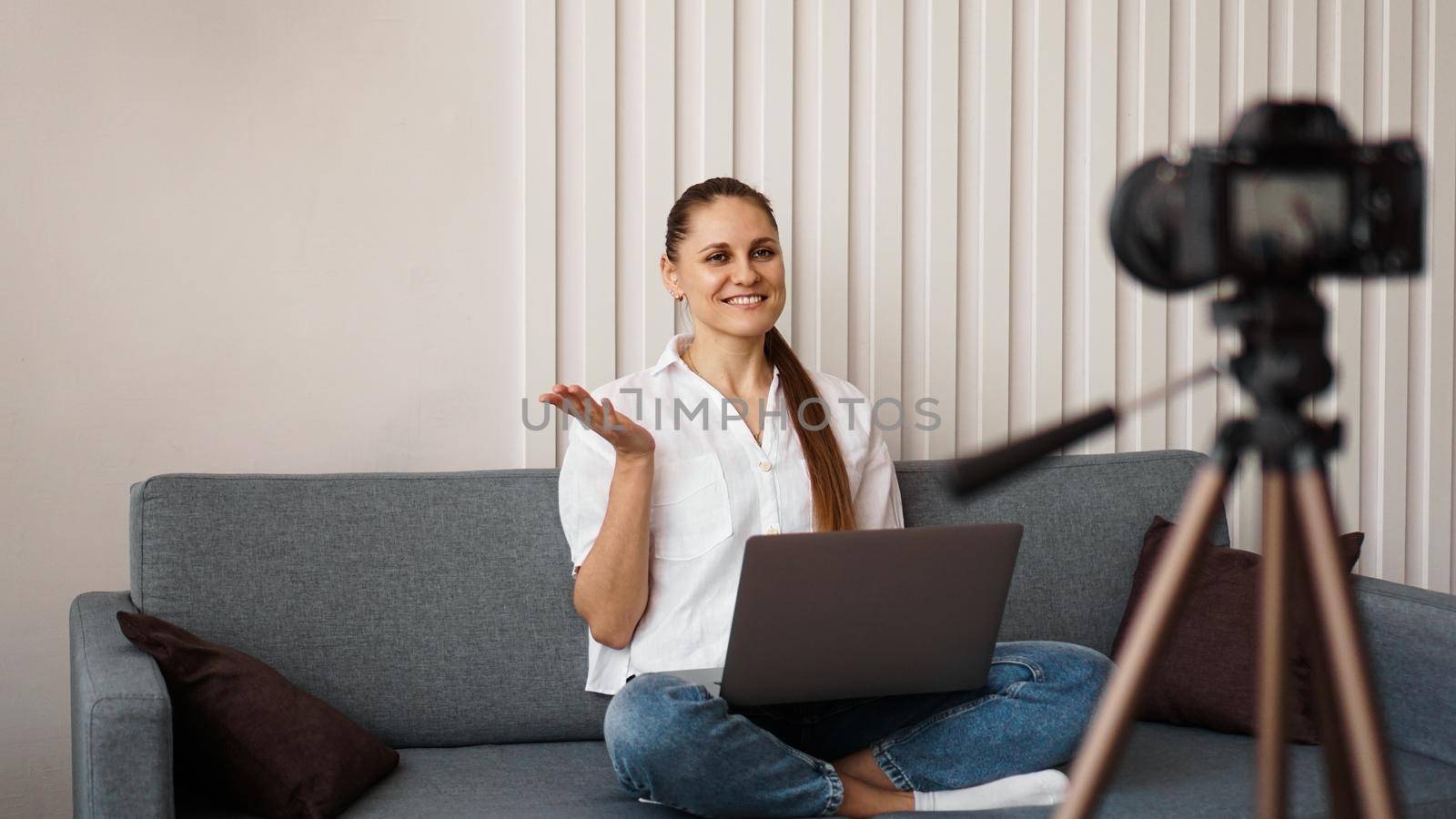 Smiling female blogger records a new video. She sits on the couch at home and holds a laptop. Positive business blog concept.