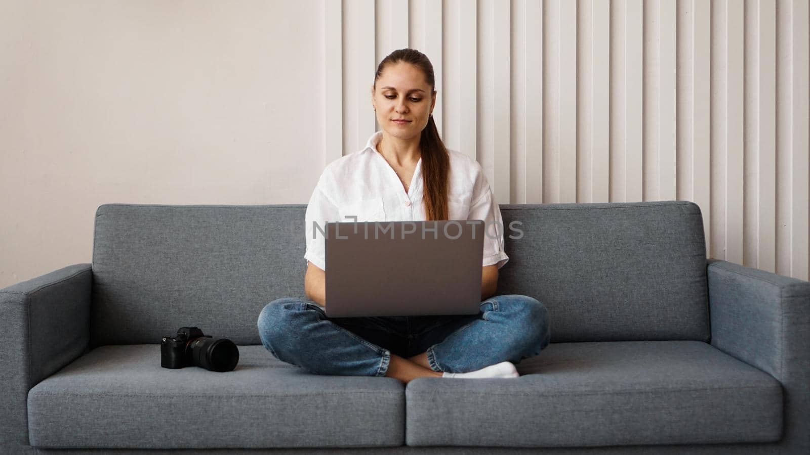 Happy young woman using laptop at home. On the couch next to the woman is a camera. Photographer retouches photographs at home.