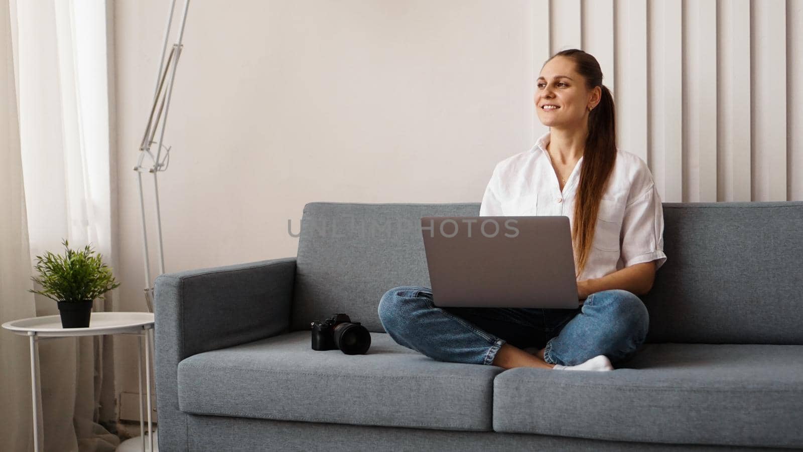 Happy young woman using laptop at home. On the couch next to the woman is a camera. Photographer retouches photographs at home.