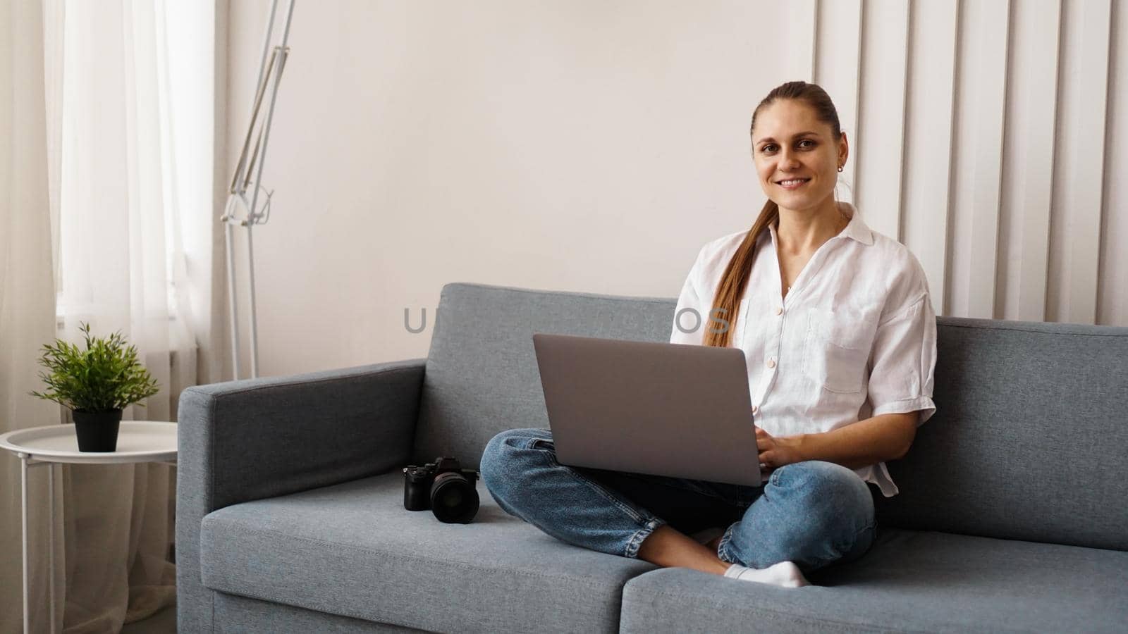 Happy young woman using laptop at home. On the couch next to the woman is a camera. Photographer retouches photographs at home.