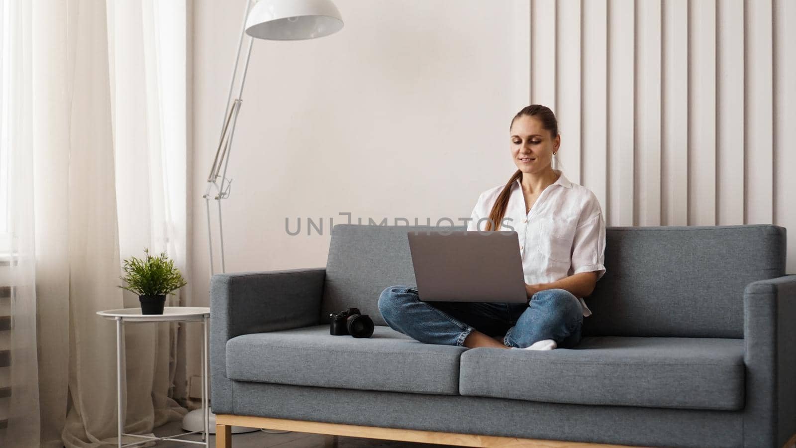 Happy young woman using laptop at home. On the couch next to the woman is a camera. Photographer retouches photographs at home.