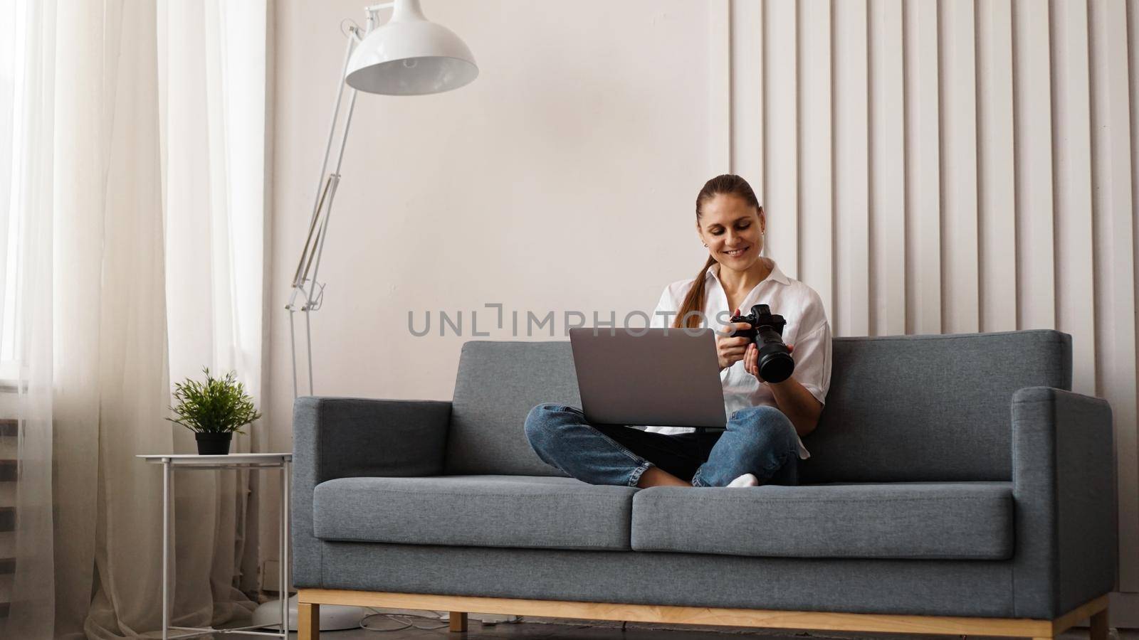 Happy young woman with photo camera using laptop at home. The photographer looks at the pictures taken and sits on the couch.