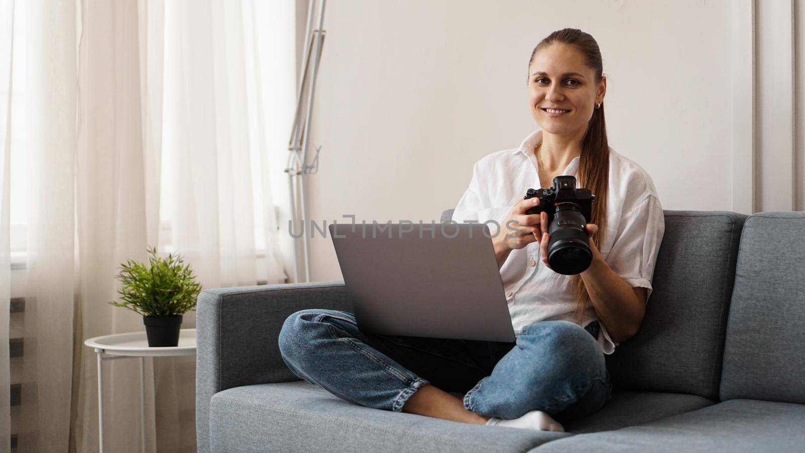 Happy young woman with photo camera using laptop at home. The photographer is holding the camera and smiling.