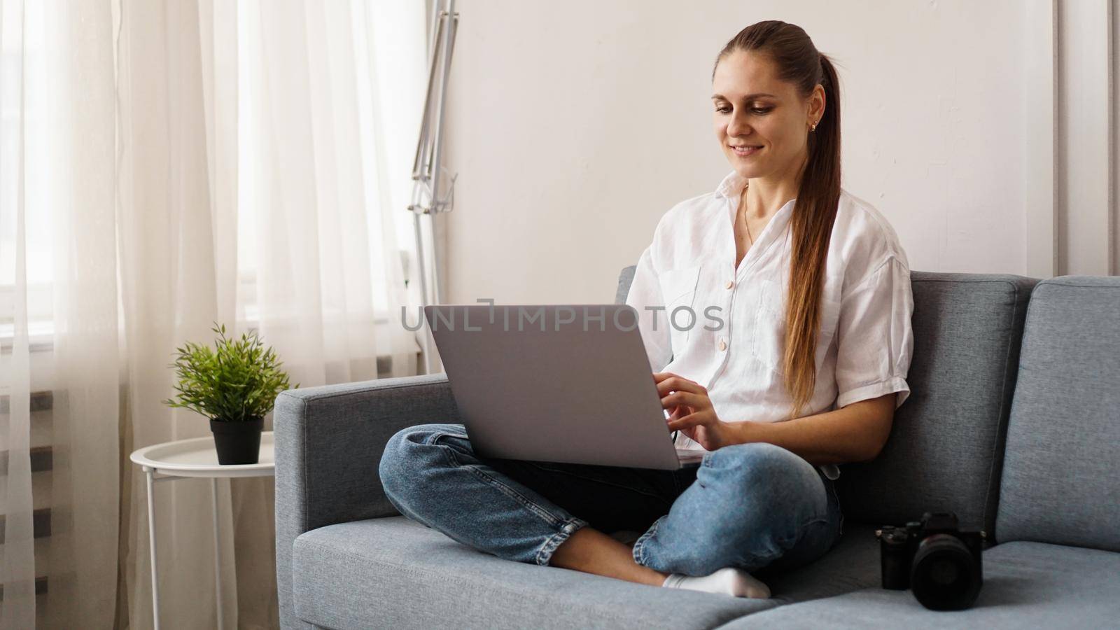 Happy young woman using laptop at home. On the couch next to the woman is a camera. Photographer retouches photographs at home.