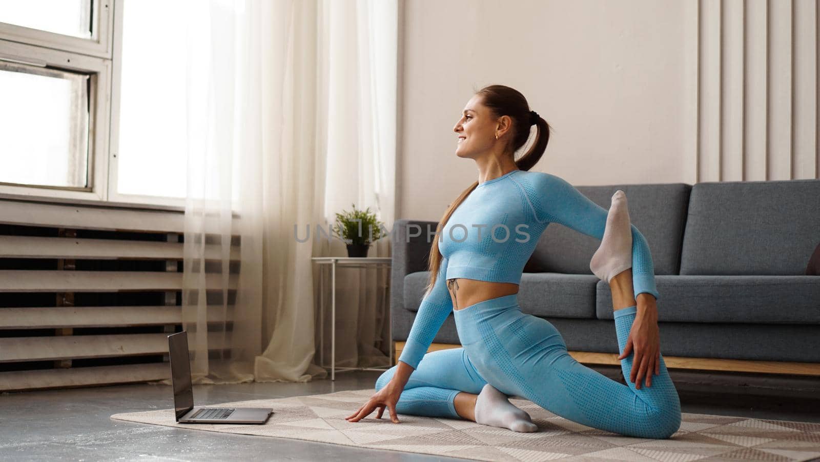 Beautiful young woman in a blue suit doing fitness exercise at home. She looks at her laptop and repeats the movements for the online course.