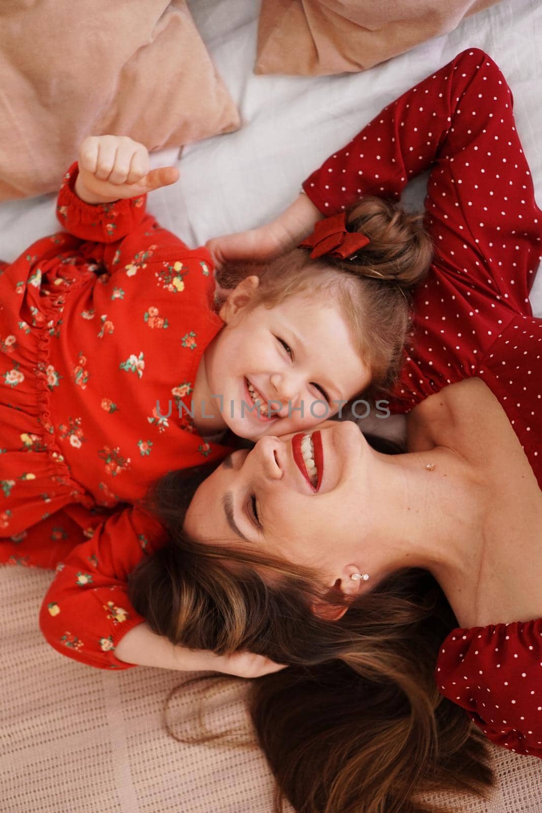 Mom and her little daughter lie on the bed facing each other. They are happy. Both are wearing red dresses.