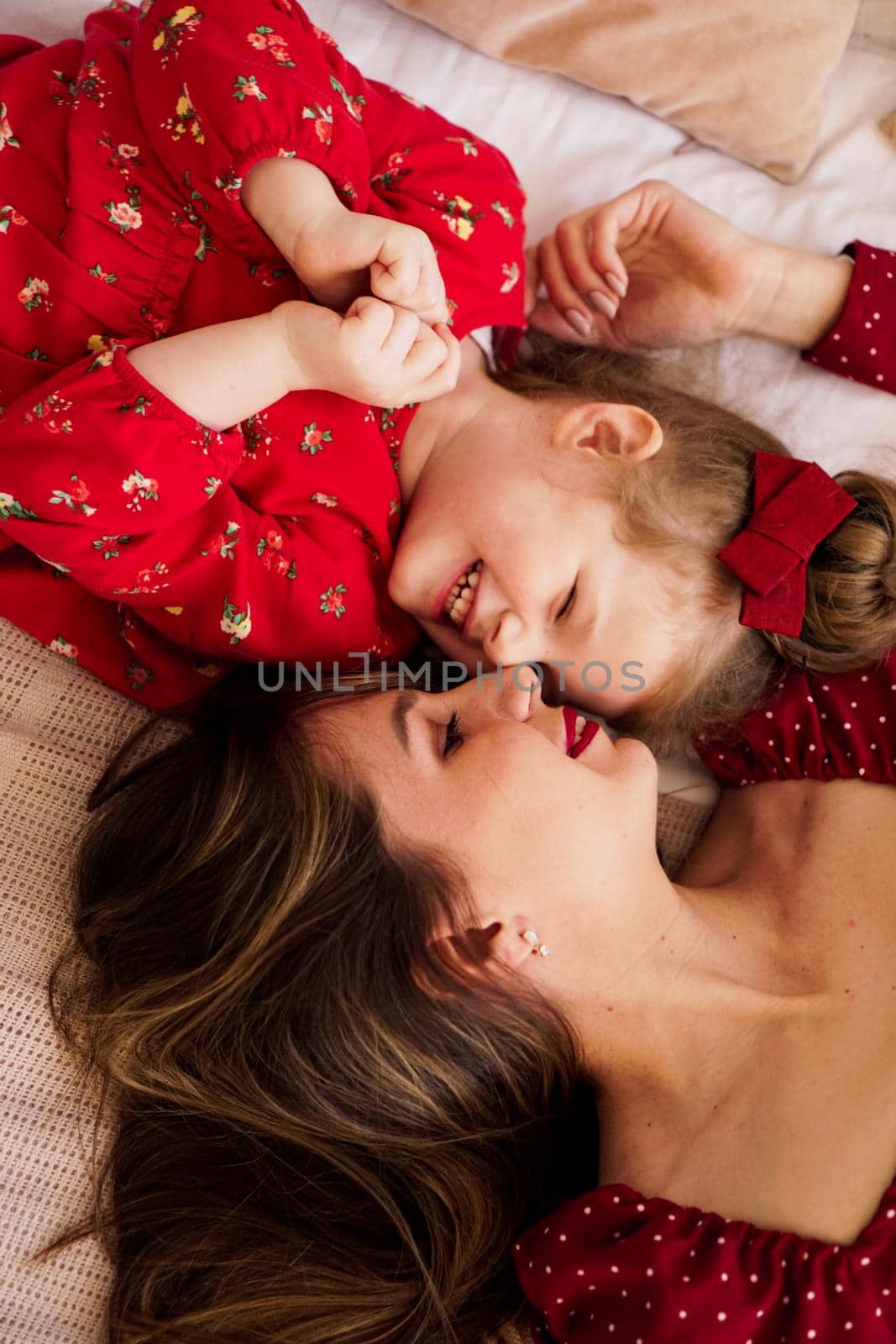 Mom and her little daughter lie on the bed facing each other. They are happy. Both are wearing red dresses.