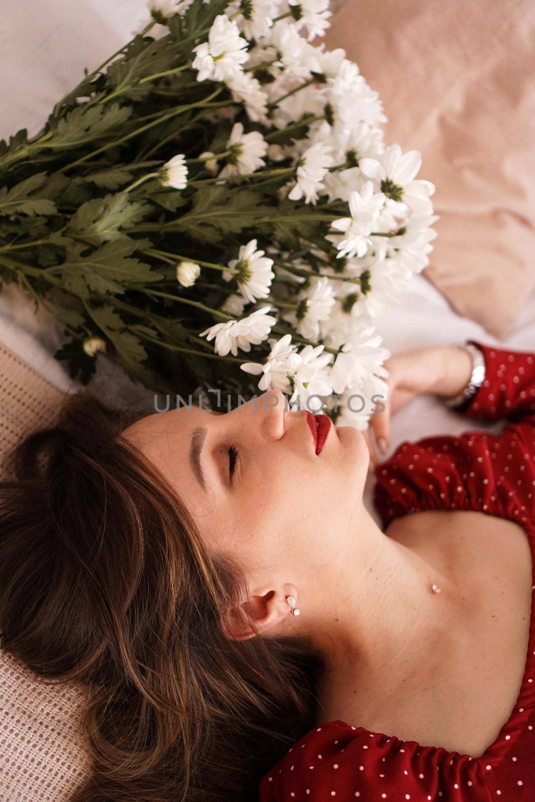 A beautiful brunette in a red dress lies on the bed next to a bouquet of daisies by natali_brill