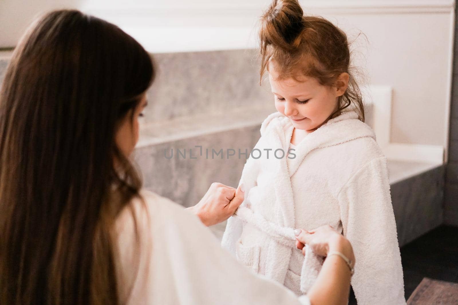 Mom puts her daughter in a white bathrobe in the bathroom. Happy family by natali_brill