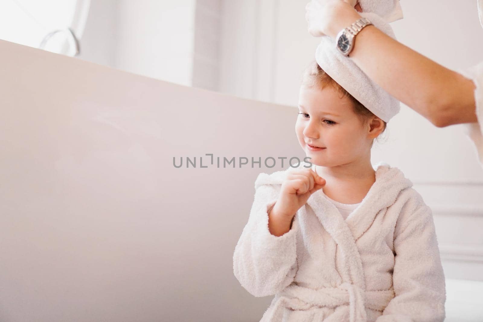 Mom puts a towel on her daughter's hair. Little daughter in a white bathrobe in a bright bathroom.