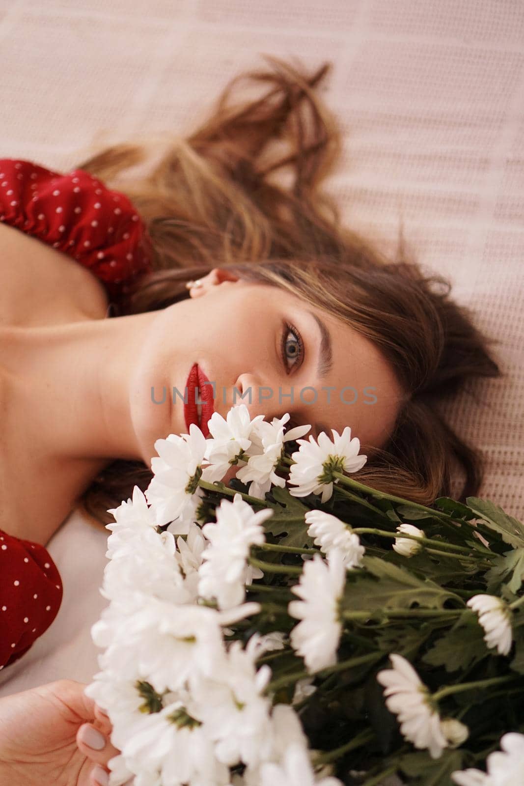 A beautiful brunette in a red dress lies on the bed next to a bouquet of daisies. Portrait of a beautiful woman.