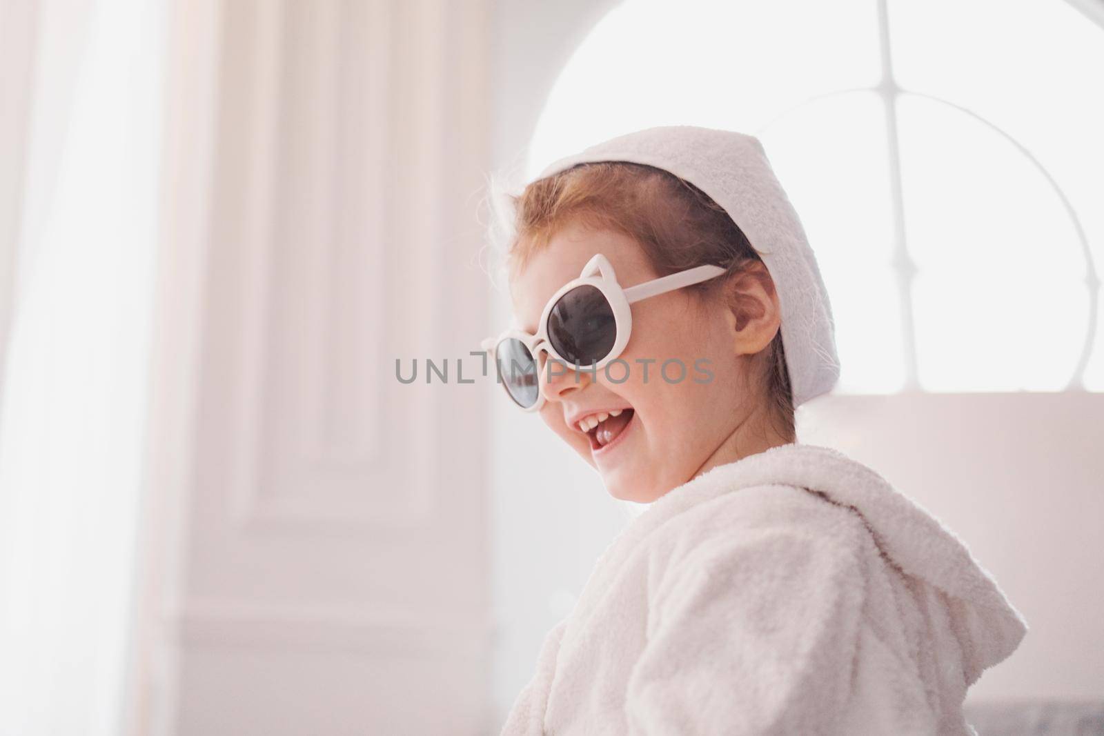 Little girl in a bathrobe and towel, sunglasses. Girl after shower in a bright interior