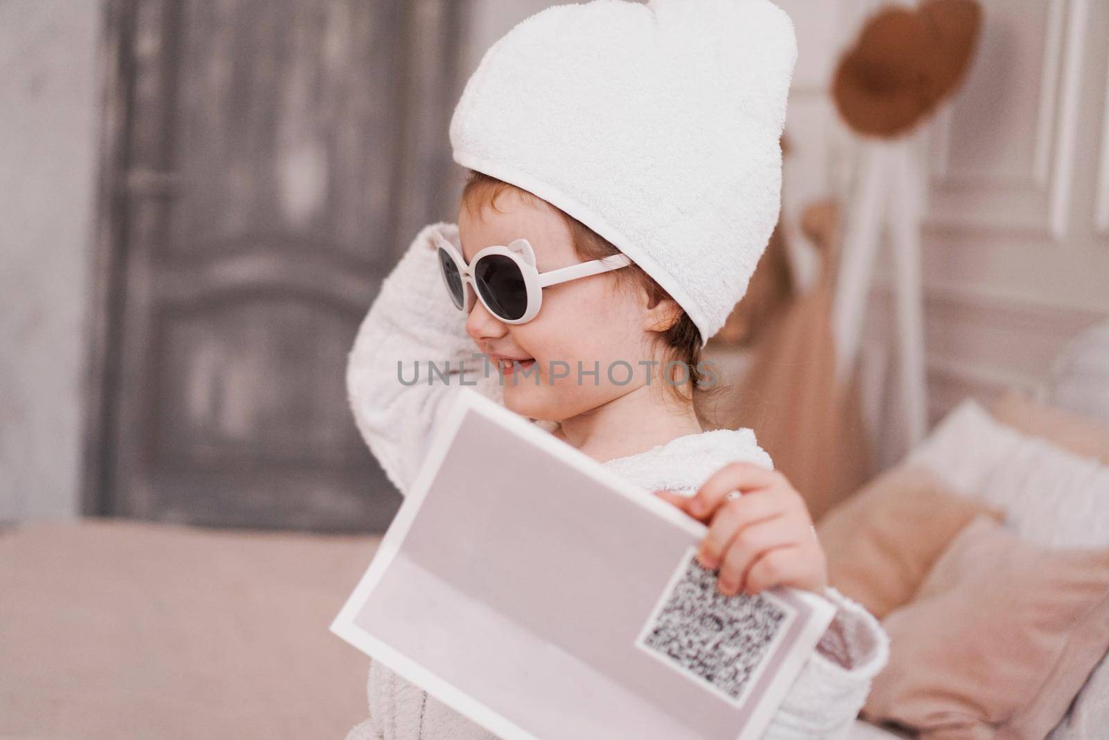Smiling little girl in a white bathrobe after a bath. White cozy interior. Hygiene and baby fashion concept. She is wearing sunglasses, the girl is holding a magazine in her hand