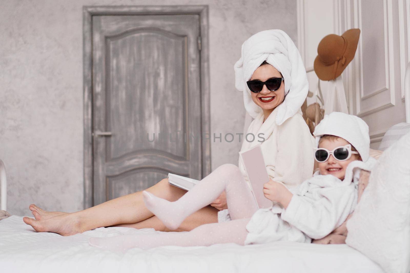 Mother and daughter in the bedroom in bathrobes. They read magazines lying on the bed. They are in sunglasses looking at the camera and laughing. Focus on mom's face