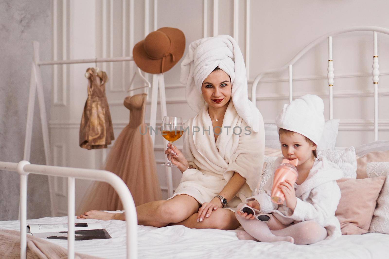 Mother and daughter in the bedroom in bathrobes. The mother drinks a glass of white wine, and the daughter drinks juice from a glass. A wonderful holiday with the family