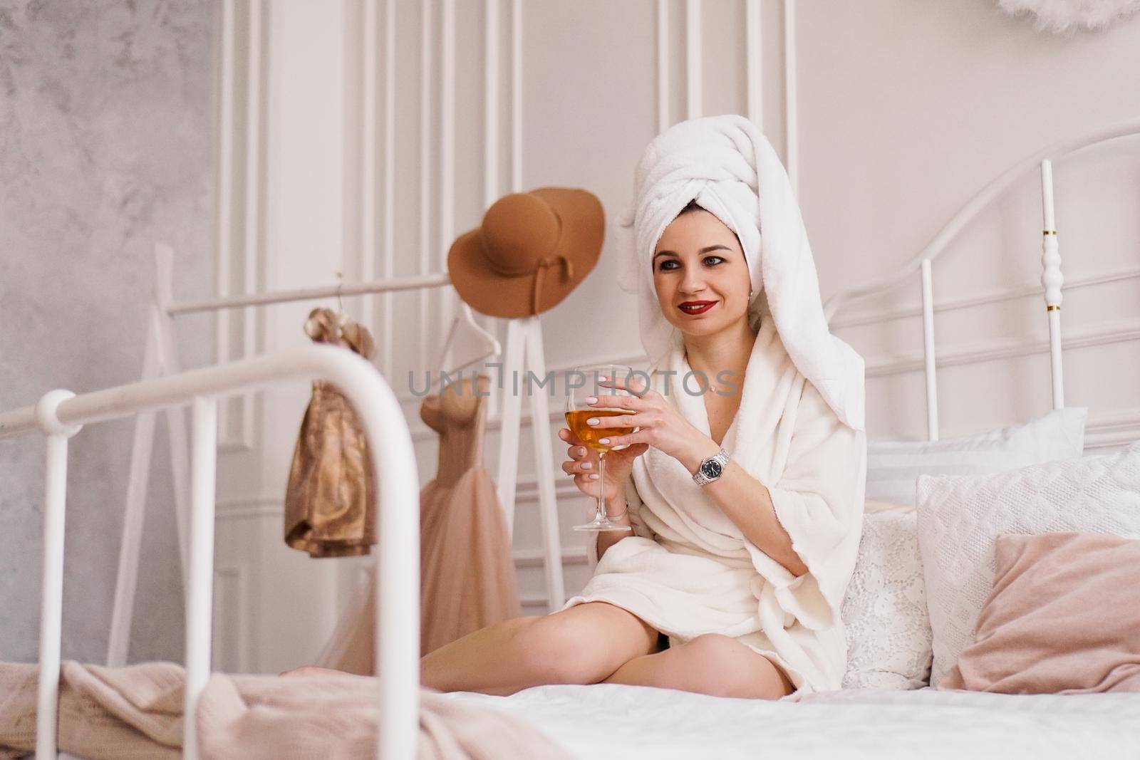 Beautiful woman in a white robe and with a towel on her hair. She is holding a glass of white wine. Light Scandinavian bedroom interior.