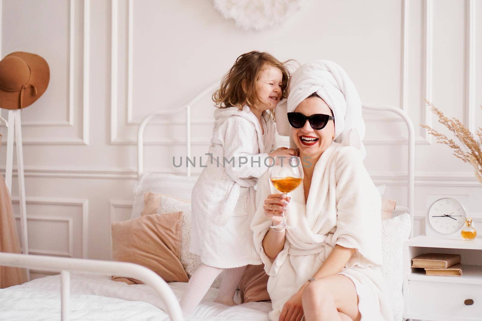 Mother and daughter in the bedroom in bathrobes. Happy daughter is jumping on the bed. Mom is drinking a glass of white wine. Funny motherhood fatigue concept