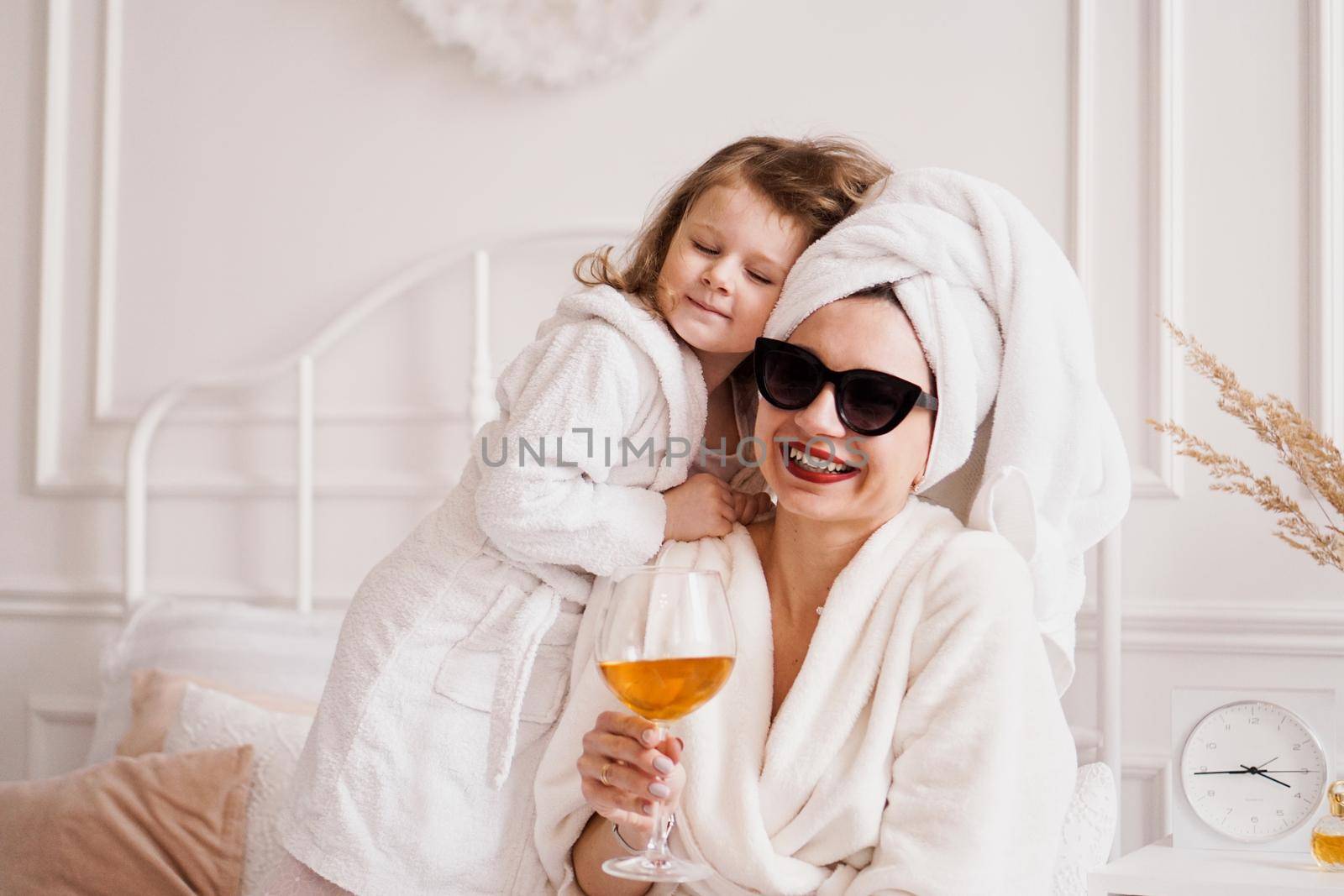 Mother and daughter in the bedroom in bathrobes. The girl hugs her mom. Mom is holding a glass of white wine.