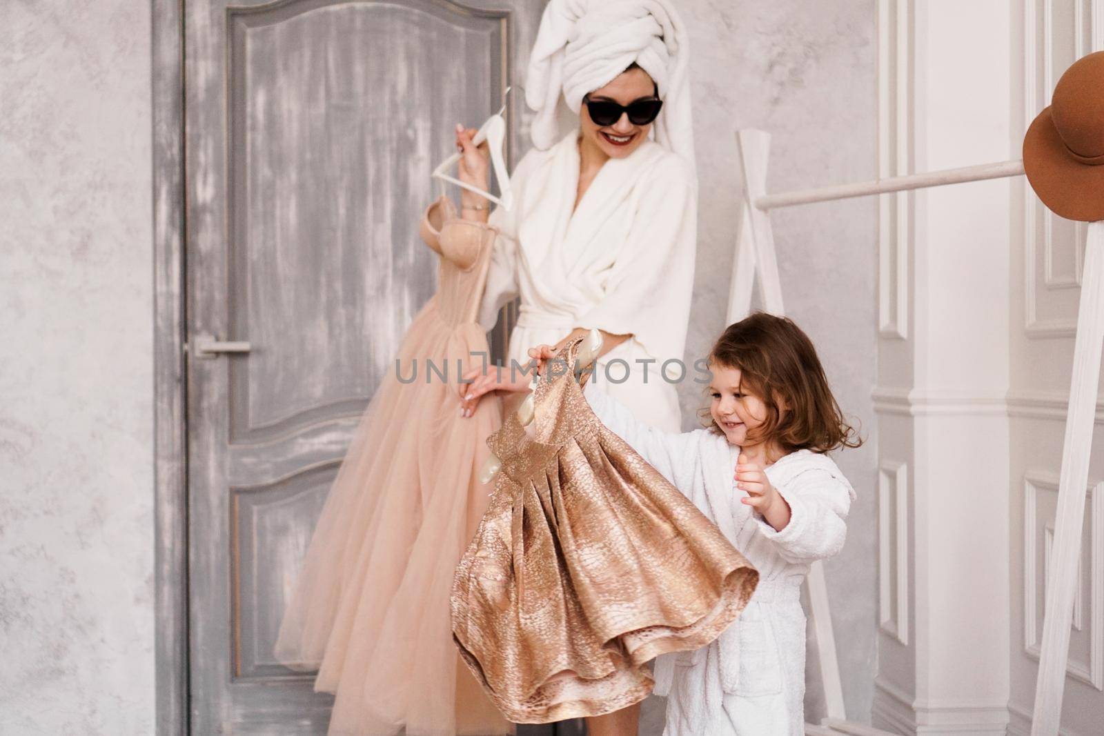 Happy mother and daughter are choosing a dress in the wardrobe. The mother is wearing a white bathrobe. They are going to the party.