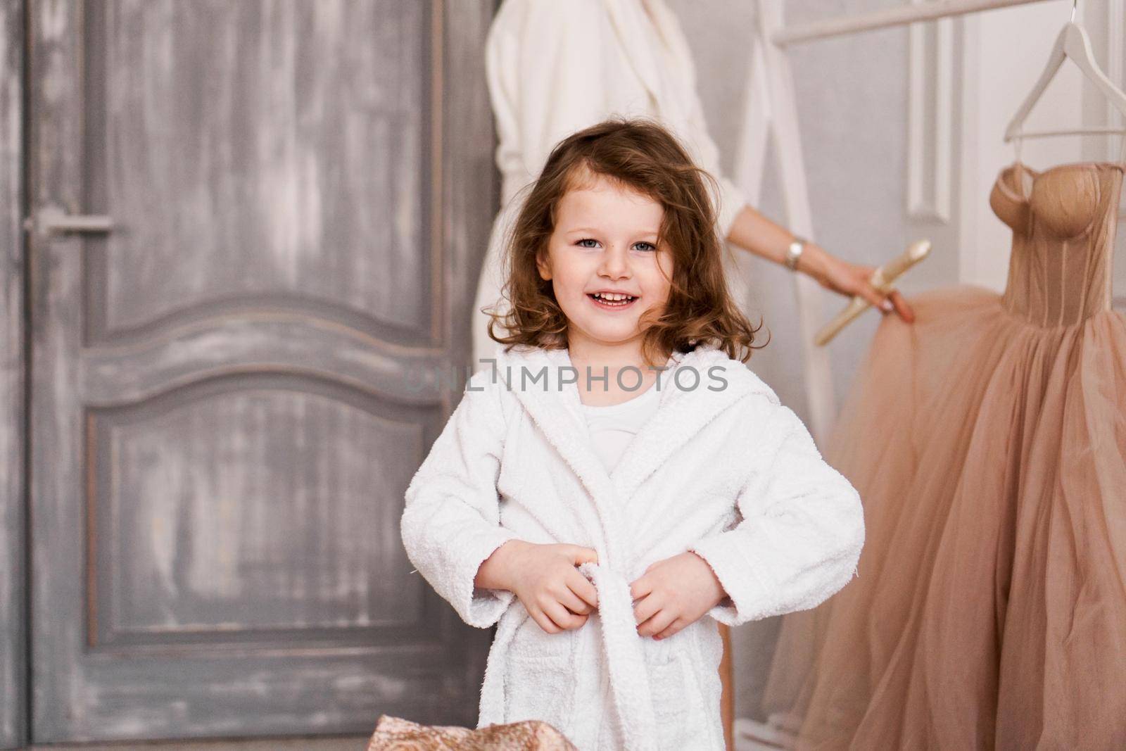 Smiling little girl in a white bathrobe after a bath. White cozy interior. Hygiene and baby fashion concept