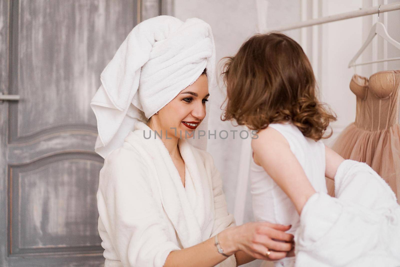 Attractive mother in a white bathrobe is dressing her little daughter. Against the background of a blurred hanger with a beige dress. Wardrobe concept