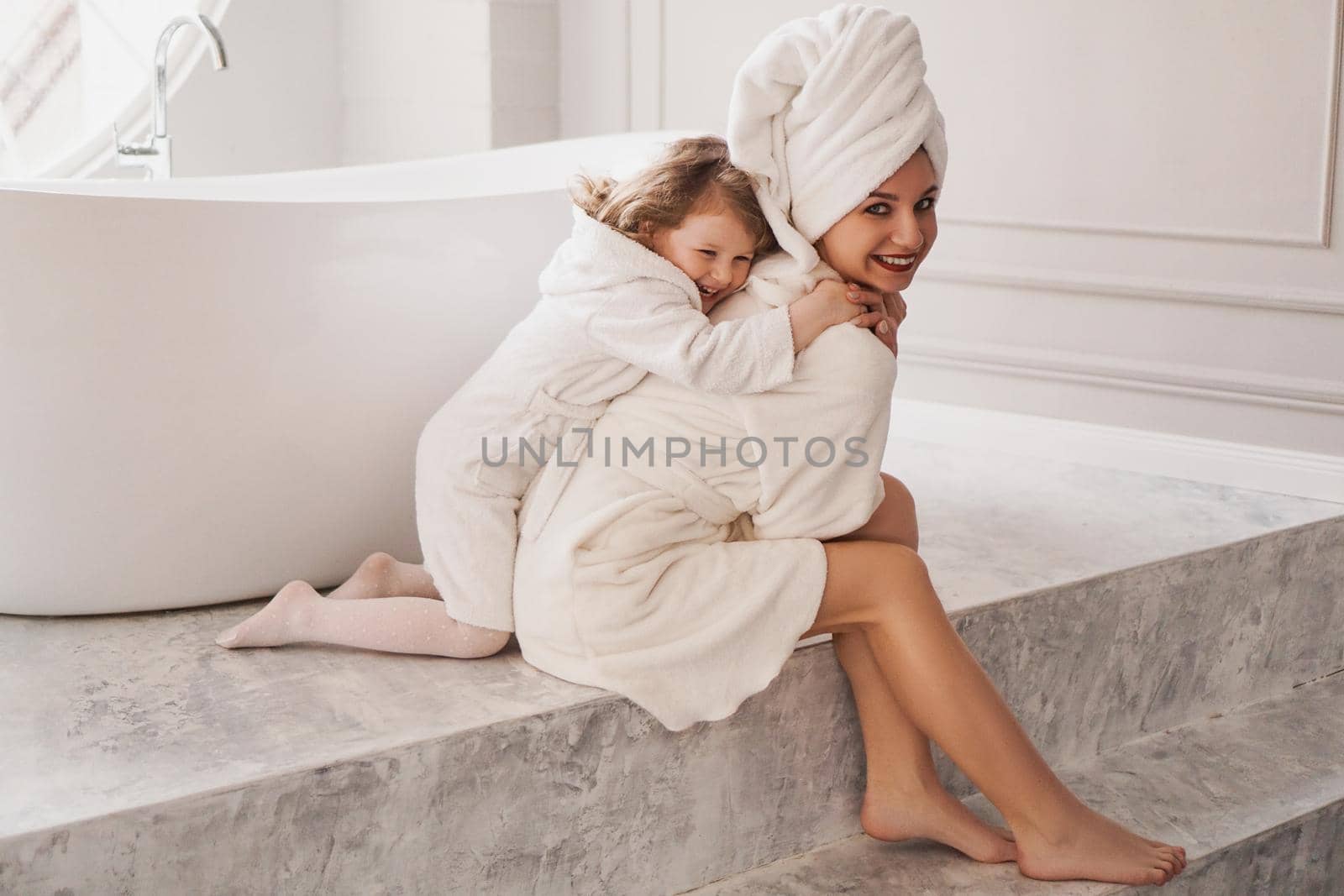 Little daughter hugs mom. Mother and daughter in white coats in a light bathroom. Hygiene, beauty and happy family concept