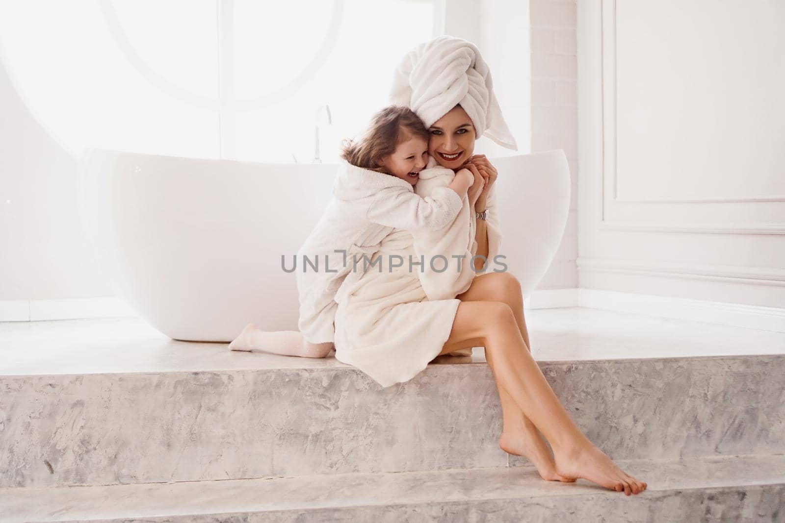 Little daughter hugs mom. Mother and daughter in white coats in a light bathroom. Hygiene, beauty and happy family concept