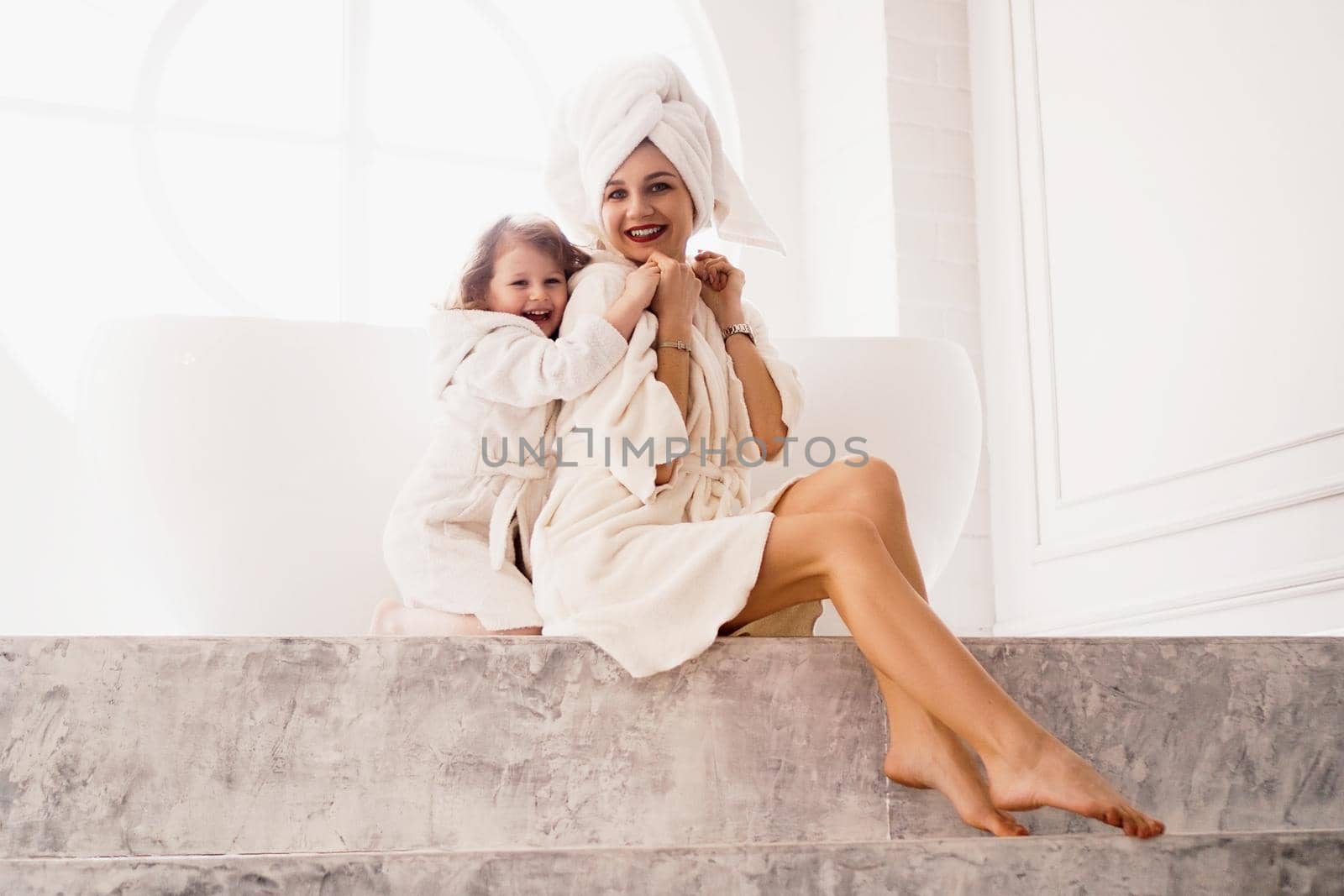 Little daughter hugs mom. Mother and daughter in white coats in light bathroom. by natali_brill