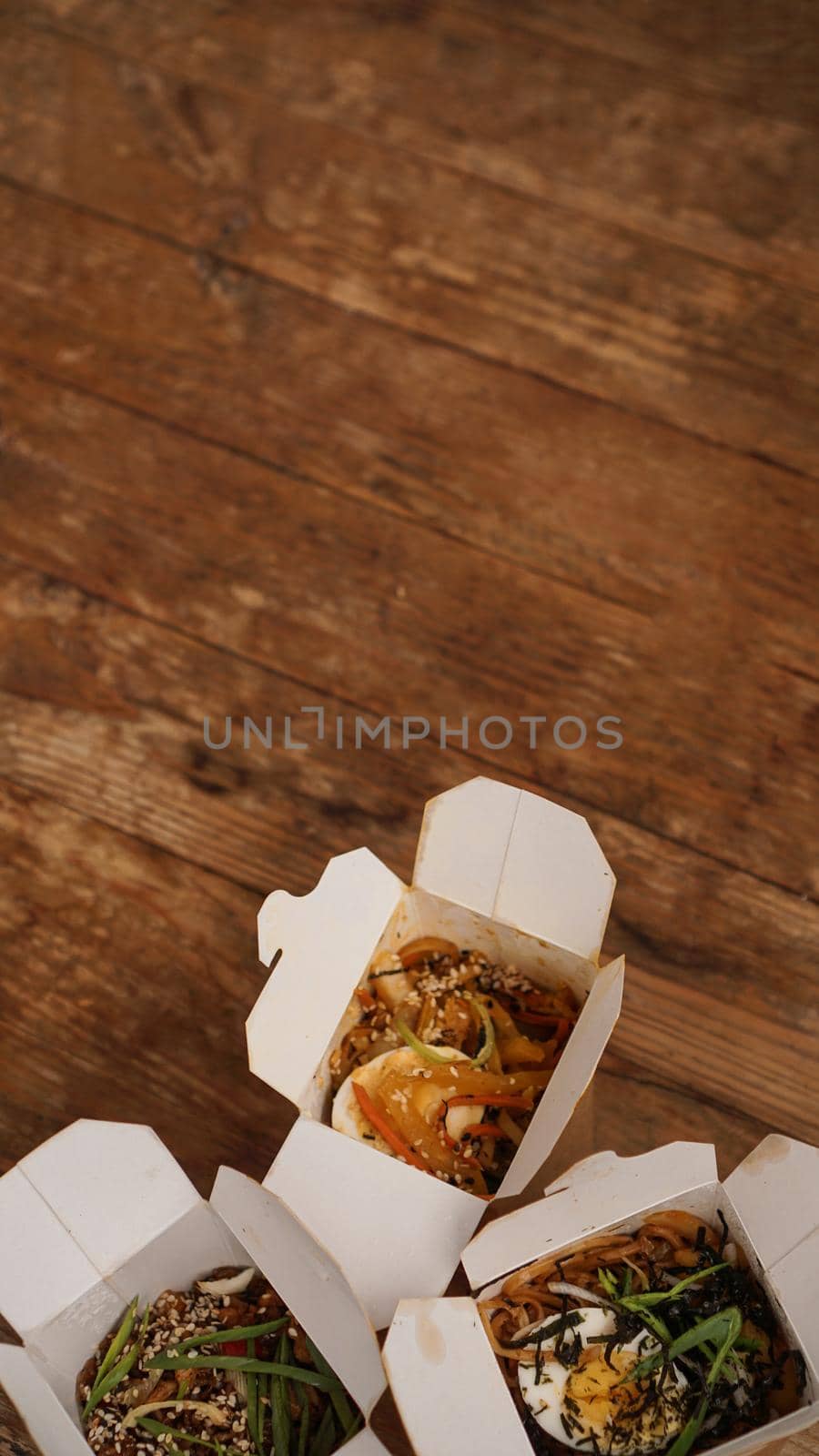 Noodles with pork and vegetables in take-out box on wooden table. Asian food delivery. Food in paper containers on wooden background. Vertical photo
