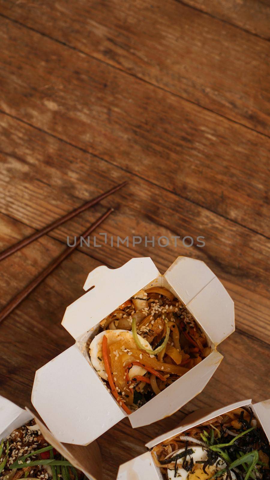 Noodles with pork and vegetables in take-out box on wooden table. Asian food delivery. Food in paper containers on wooden background. Vertical photo