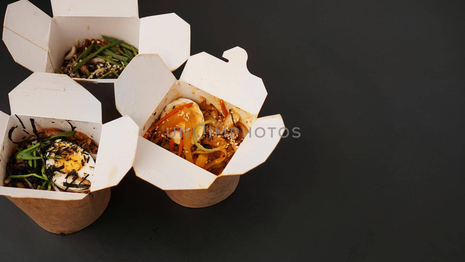 Noodles with pork and vegetables in take-out box on black table. Asian food delivery. Food in paper containers on black background