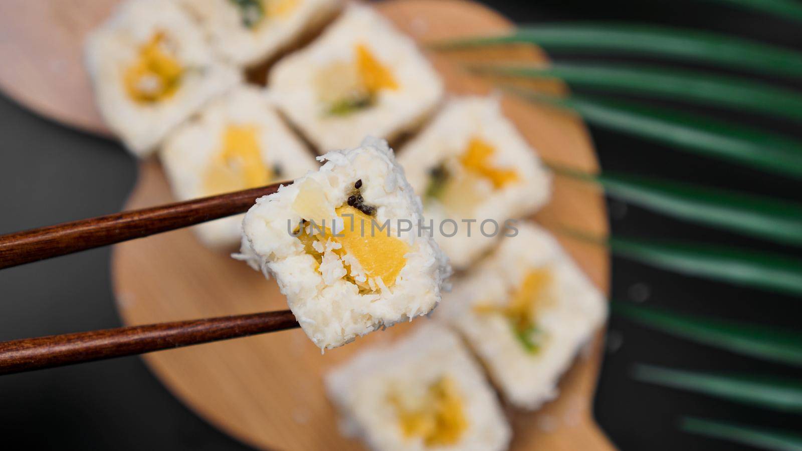 Dessert sushi. Sweet kiwi, pineapple sushi rolls. Sushi on a wooden tray on a black background. Holding a sweet roll with wooden sticks.