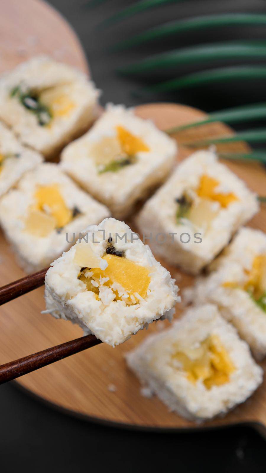 Dessert sushi. Sweet kiwi, pineapple sushi rolls. Sushi on a wooden tray on a black background with a tropical leaf. Vertical photo