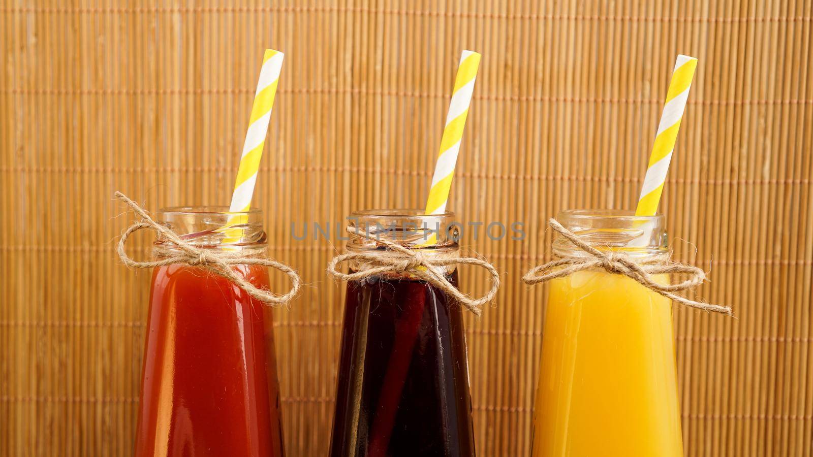 Three multicolored drinks in the bottles on a wooden bamboo background. Bottled paper straws. The bottle is tied with twine. Homemade drink